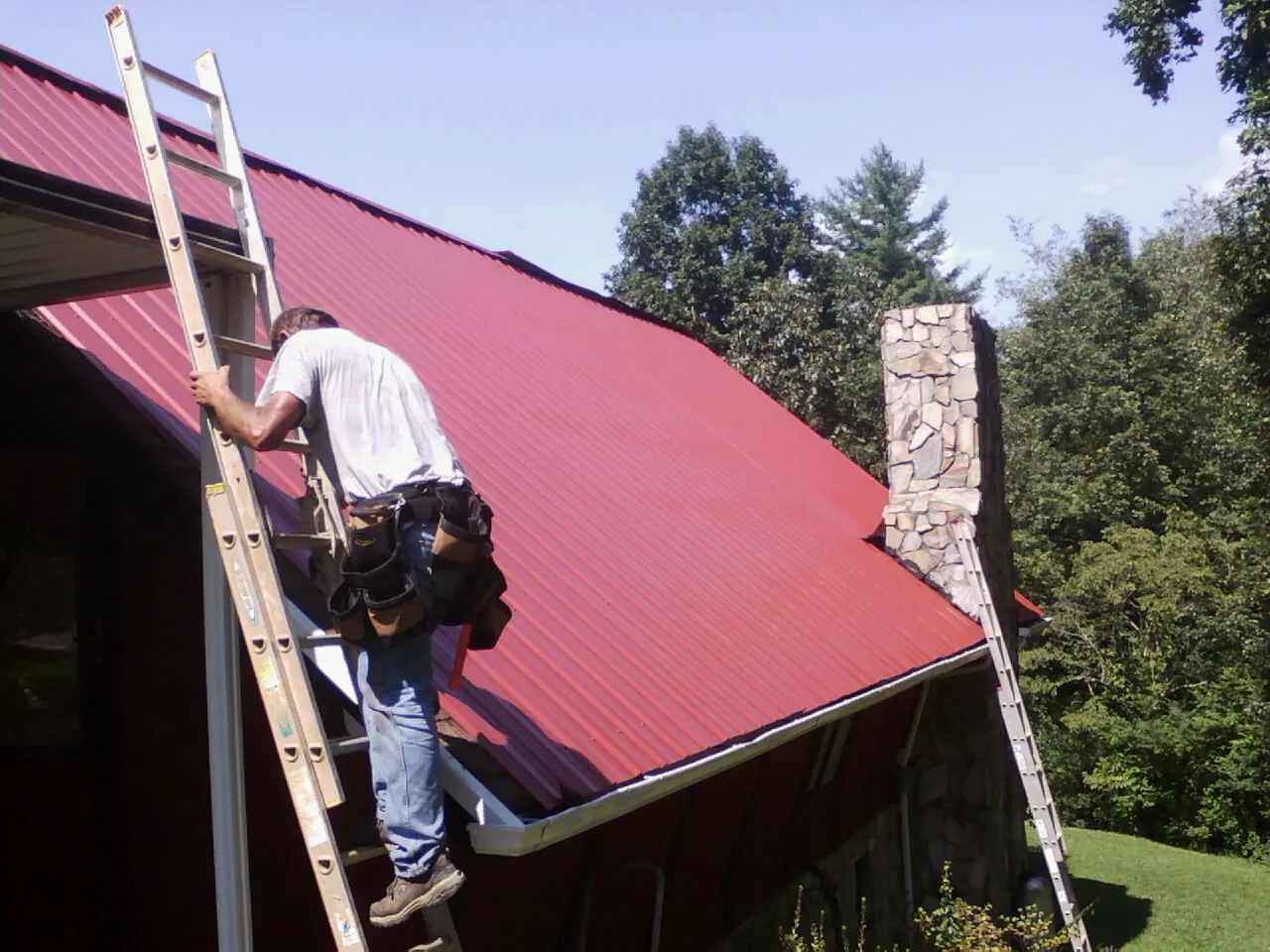 Roofer on a ladder beside a red metal roof and stone chimney — Soffit & Fascia Repair by Roofing Brothers in Tallapoosa